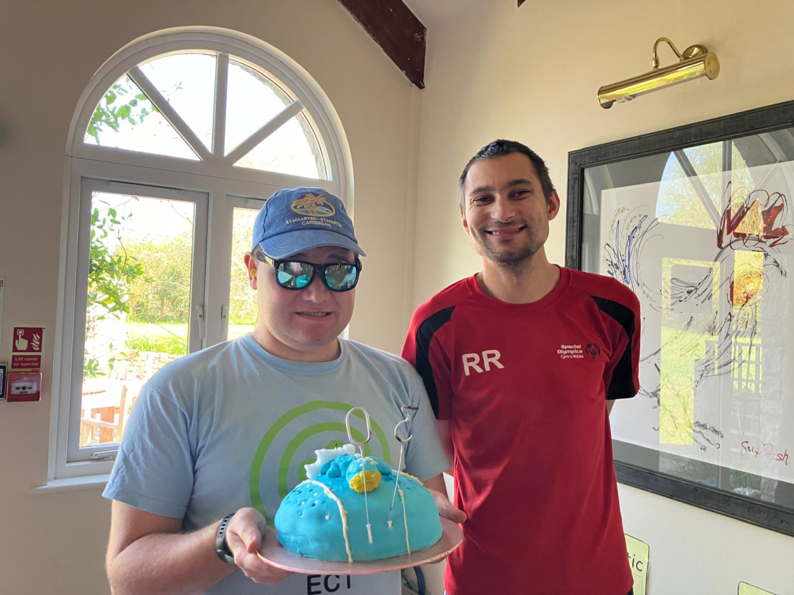 Two young men carry a birthday cake. One has a cap and sunglasses and one wears a red shirt. The cake is blue.