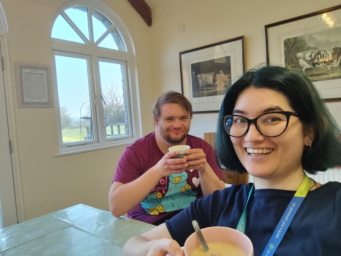 A smiling woman in glasses with a hot drink. A young man in the background with a hot drink. A picture window behind.