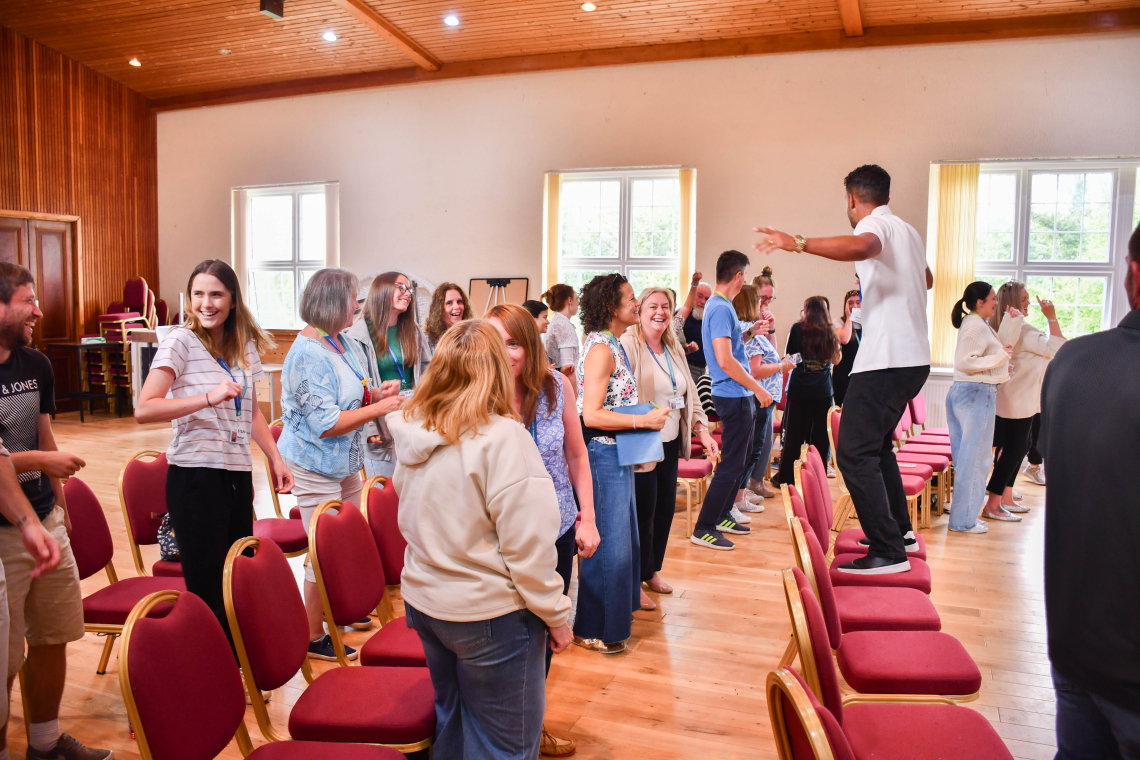 A room full of people dance in front of chairs lined up in rows in a hall. A man stands on a chair in front of them