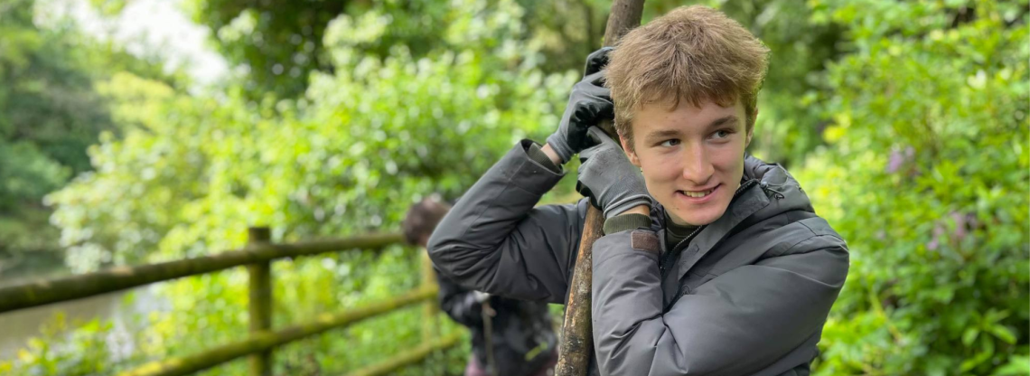 A boy leans on a stick. He is in a wood. He smiles