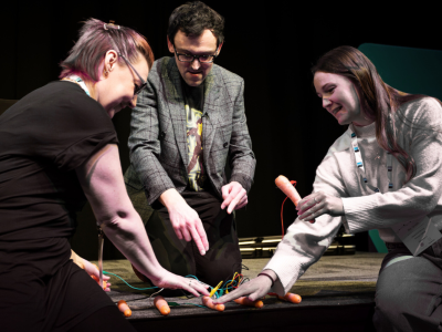 Three people on a stage use carrots to play music