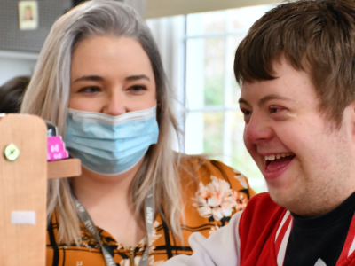 Support worker wearing a mask with a young person laughing at a weaving loom.