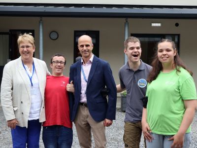 5 people smile in a line. There is a woman in a cream jacket and blue lanyard. A young man in a red top. A man in a navy jacket. A young man and woman in t-shirt