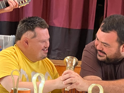 Two men hold a brass handbell. They sit in front of a stage