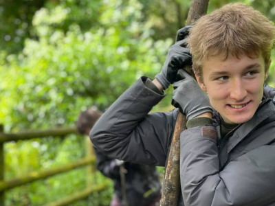 A boy leans on a stick. He is in a wood. He smiles