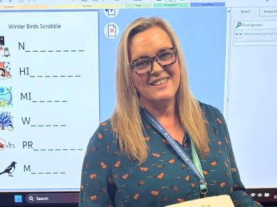 A female tutor with blonde hair and classes stands in front of a white board. She is smiling.